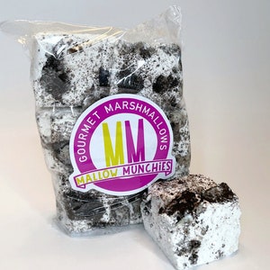 May include: A clear plastic bag filled with gourmet marshmallows. The marshmallows are white with dark cookie pieces throughout. A single marshmallow sits outside the bag. The bag has a pink and yellow label that reads "GOURMET MARSHMALLOWS MM MALLOW MUNCHIES."