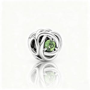 May include: A silver charm with a woven pattern, showcasing a round, light green gemstone in the middle. The charm is highly reflective, with a clear reflection beneath. The design is intricate and detailed, suitable for a bracelet or necklace.
