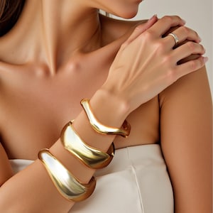 May include: Three gold-toned metal bangles stacked on a person's wrist. The bangles are all different sizes and have a wavy, organic shape.