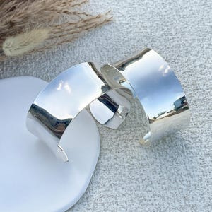 May include: Two silver bangle bracelets, one slightly larger than the other, are stacked on top of each other. The bracelets are shiny and smooth.