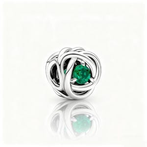 May include: A silver charm with a complex, interwoven pattern. It has a bright, round, emerald-green gemstone at its core. The reflective surface below mirrors the charm's shape, creating a symmetrical effect.