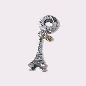 Puede incluir: Charm de plata con un diseño detallado de la Torre Eiffel. El charm tiene una parte superior circular con la inscripción "PANDORA" y "ALE S925". Se adjunta un pequeño acento dorado.