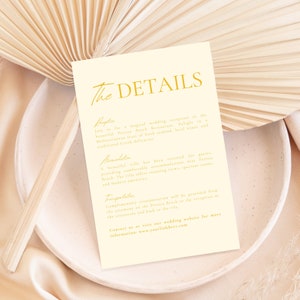 May include: A cream-colored card with gold text detailing the reception location, accommodation, and transportation for a wedding. The text reads "The Details", "Reception", "Join us for a magical wedding reception in the beautiful Perissa Beach Restaurant. Delight in a Mediterranean feast of fresh seafood, local wines, and traditional Greek delicacies.", "Accommodation", "A beautiful villa has been reserved for guests providing comfortable accommodations near Perissa Beach. The villa offers stunning views, spacious rooms, and modern amenities.", "Transportation", "Complimentary transportation will be provided from the ceremony on the Perissa Beach to the reception in the restaurant and back to the villa.", "Contact us or visit our wedding website for more information: www.yourlinkhere.com"