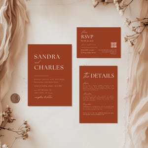 May include: A set of terracotta-colored wedding invitation cards. The main card features the names "Sandra and Charles" in white, with event details. Additional cards include RSVP and event details. Floral and fabric accents surround the cards.