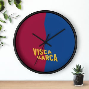 May include: A circular wall clock with a black surround. The clock face is divided into burgundy and blue halves. The words "VISCA BARCA" are printed in yellow. The clock hands are black.