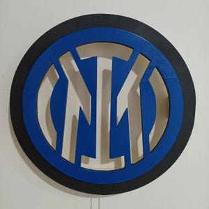 Könnte beinhalten: Eine Holz-Wanddekoration mit dem Logo von Inter Mailand. Das Logo ist ein kreisförmiges Design mit einem blauen und schwarzen Rand und einem blauen Innenkreis mit den Buchstaben "IM" in Weiß. Das Logo ist an einer weißen Wand befestigt.