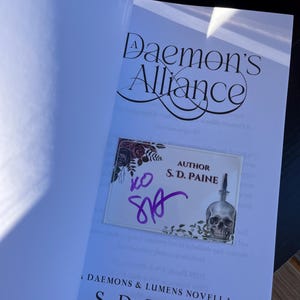 May include: A white book with the title "A Daemon's Alliance" and the author's name "S.D. Paine" printed on the cover. The book is signed by the author with a purple signature.