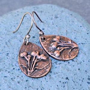 May include: A pair of copper-colored teardrop earrings. Each earring features a floral design with detailed leaves and buds. The earrings have silver-colored ear wires and a textured finish, showcasing a rustic aesthetic.