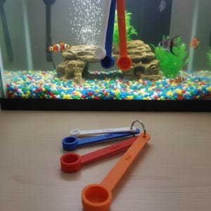 May include: A set of colorful measuring spoons in white, blue, red, and orange, attached to a metal ring. The spoons are labeled with measurements. An aquarium with fish and colorful gravel is in the background.