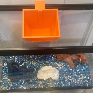 May include: An orange plastic fish food dispenser hanging above a fish tank. The tank is filled with blue and white gravel and contains a piece of driftwood and a white paper object.