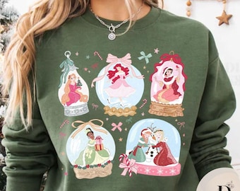 Könnte beinhalten: Grünes Sweatshirt mit sechs Schneekugeln, die Prinzessinnen-Illustrationen zeigen. Jede Kugel hat eine andere Prinzessin, mit Zuckerstangen und Schleifen. Das Sweatshirt ist einfarbig grün, perfekt für die Weihnachtszeit.