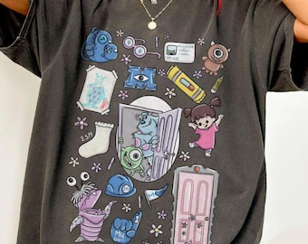 May include: Dark gray t-shirt featuring a colorful graphic of characters and elements from the animated film Monsters, Inc. The design includes Mike Wazowski, Sully, Boo, and other recognizable items. The shirt has a relaxed fit.