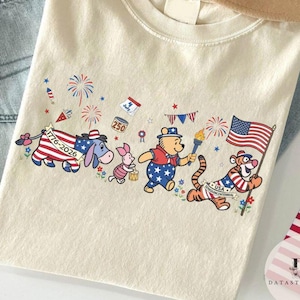 Op de afbeelding: Cr&egrave;mekleurig T-shirt met een patriottisch ontwerp met stripfiguren. Het ontwerp bevat Winnie de Poeh, Knorretje, Iejoor en Tijger, elk versierd met kleding in Amerikaanse vlagstijl en met patriottische symbolen.