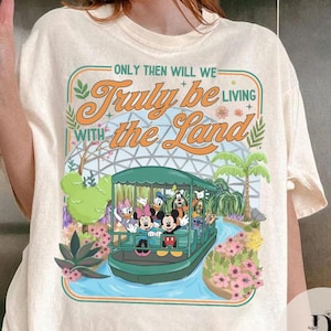 May include: Cream-colored t-shirt featuring a colorful graphic of Disney characters on a boat ride. The design includes Mickey, Minnie, Donald, Daisy, Goofy, and other characters. The text reads "Only Then Will We Truly Be Living With the Land."