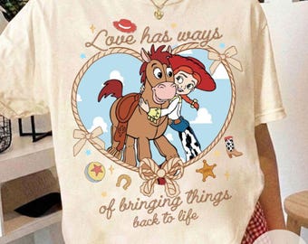 May include: A cream-colored t-shirt featuring a graphic of Jessie and Bullseye from Toy Story inside a heart-shaped rope frame. The text reads Love has ways of bringing things back to life, surrounded by small icons like a cowboy hat and star.