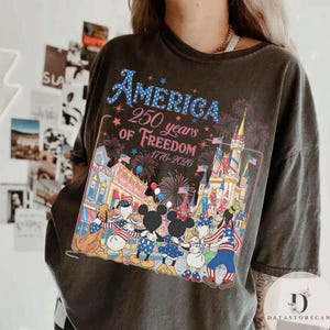 May include: Dark grey t-shirt with a vibrant graphic commemorating 250 years of American freedom. The design incorporates fireworks, a castle, and cartoon characters. The text "AMERICA 250 years of Freedom 1776-2026" is displayed in blue.