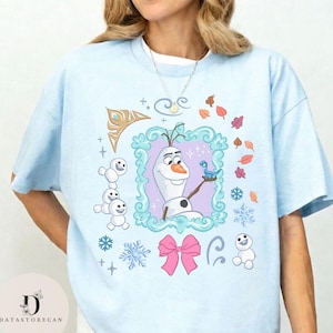 Peut inclure: T-shirt bleu clair avec un motif d'Olaf de La Reine des Neiges, encadr&eacute; dans une bordure d&eacute;corative. Le motif comprend des flocons de neige, des feuilles et un n&oelig;ud rose. Le t-shirt a des manches courtes et un col rond.