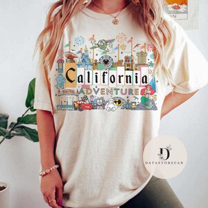 Op de afbeelding: Een wit T-shirt met een kleurrijk grafisch ontwerp met de woorden "California Adventure" en verschillende Disney-figuren, waaronder Mickey Mouse, Ariel en Buzz Lightyear.