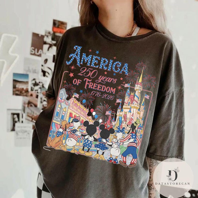 May include: Dark grey t-shirt with a vibrant graphic commemorating 250 years of American freedom. The design incorporates fireworks, a castle, and cartoon characters. The text "AMERICA 250 years of Freedom 1776-2026" is displayed in blue.