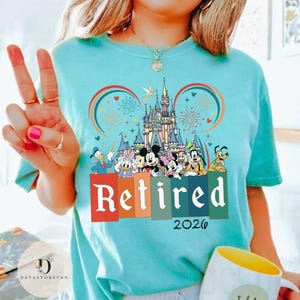 May include: A light blue t-shirt featuring a colorful graphic of Disney characters in front of a castle, with the word "Retired" and the year "2026". The design includes Mickey and Minnie Mouse, Donald Duck, and Goofy.