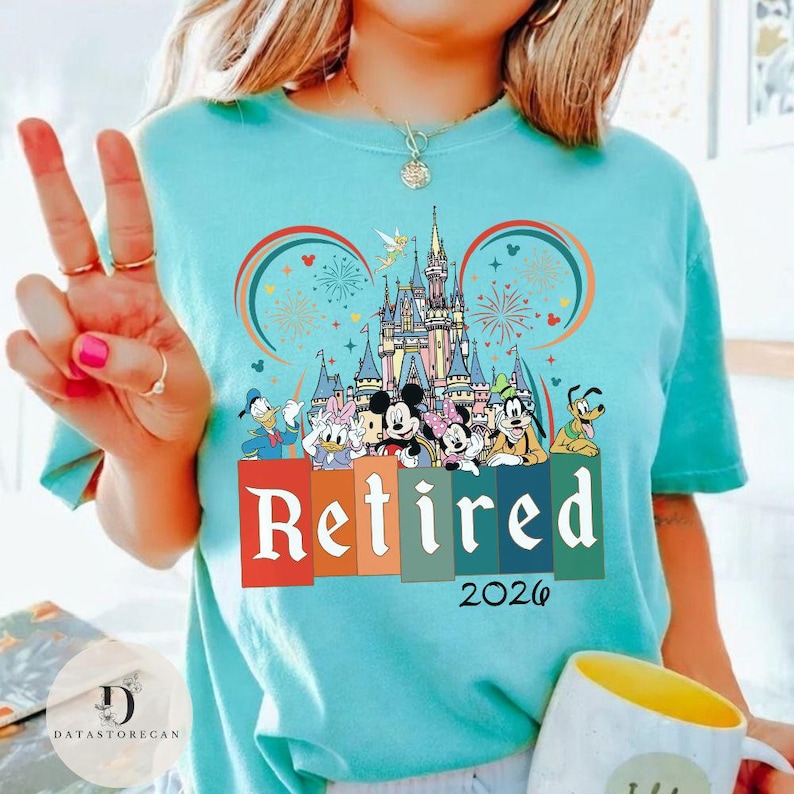 May include: A light blue t-shirt featuring a colorful graphic of Disney characters in front of a castle, with the word "Retired" and the year "2026". The design includes Mickey and Minnie Mouse, Donald Duck, and Goofy.