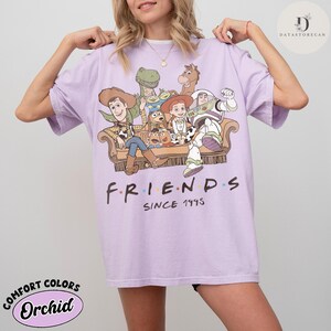 May include: A light purple short-sleeve t-shirt featuring a graphic of Toy Story characters sitting on a couch. The text below the graphic reads FRIENDS SINCE 1995. The shirt has a crew neckline and a relaxed fit, showcasing a colorful illustration.