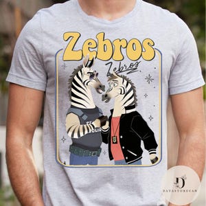 Pu&ograve; includere: T-shirt grigio chiaro con un design a cartoni animati di due zebre. Il testo giallo dice "Zebros". Una zebra indossa occhiali da sole e un giubbotto della polizia. L'altra zebra indossa una giacca nera. Il design &egrave; incorniciato.