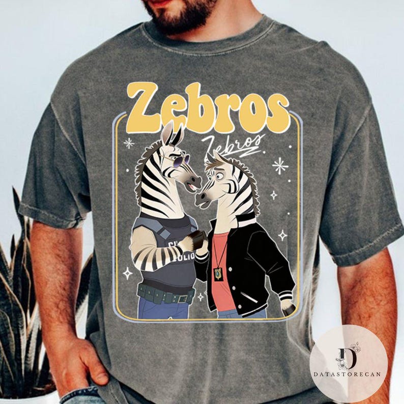 Pu&ograve; includere: T-shirt grigia con un design a cartoni animati di due zebre. Una zebra indossa occhiali da sole e un giubbotto della polizia, l'altra una giacca nera. Il testo "Zebros" &egrave; in un rettangolo giallo.
