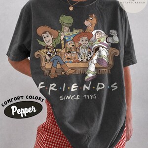 May include: A charcoal gray t-shirt featuring a graphic print of Toy Story characters sitting on a couch. The text reads FRIENDS SINCE 1995. The shirt has a crew neckline and short sleeves. The graphic includes Woody, Buzz Lightyear, Jessie, Rex, Bullseye, Slinky Dog, and Mr. Potato Head in a vintage-style illustration.