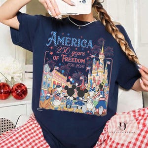 May include: Navy blue t-shirt with the text "America 250 years of Freedom 1776-2026". The graphic features a patriotic design with fireworks, a castle, and cartoon characters. The shirt is a celebration of American history.
