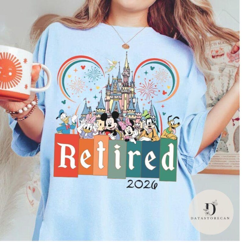 May include: Light blue t-shirt featuring a colorful graphic of Disney characters in front of a castle, with the word "Retired" and the year "2026". The design includes Mickey and Minnie Mouse, Donald Duck, and Goofy.