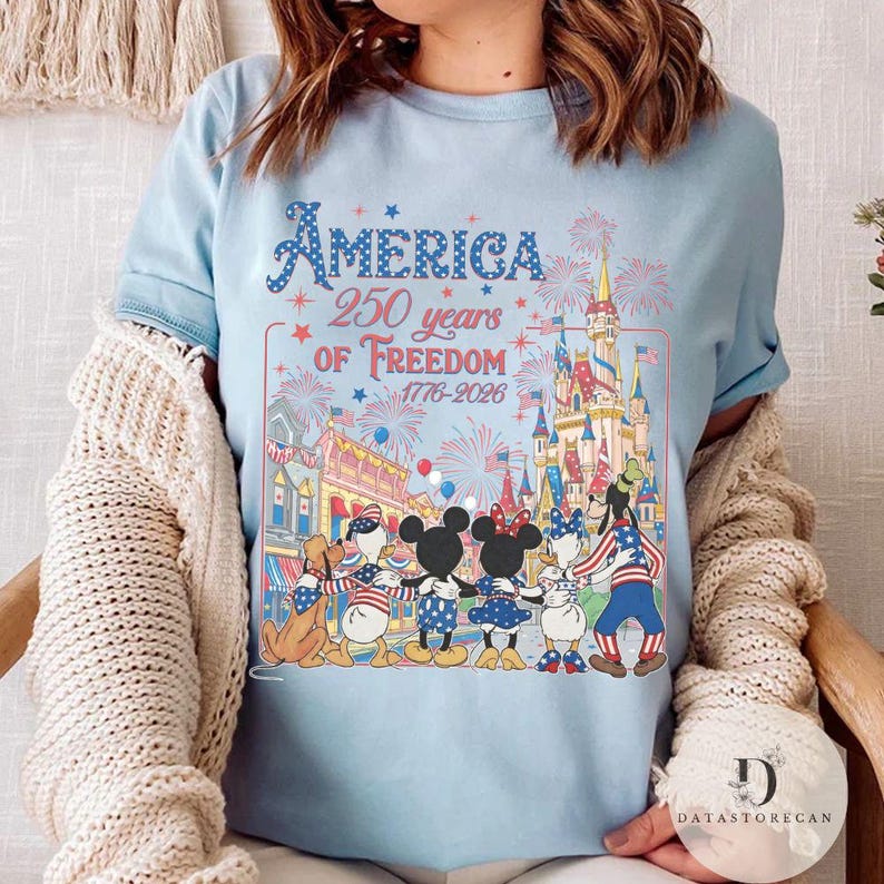 May include: Pale blue t-shirt with the text "America 250 years of Freedom 1776-2026". The shirt features a graphic of Disney characters in patriotic attire in front of a castle and fireworks.