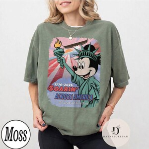 May include: Sage green t-shirt with a graphic of Mickey Mouse as the Statue of Liberty, holding a torch. The design includes the text "1776-2026 Soarin' Across America" with a patriotic red, white, and blue background. The shirt has a relaxed fit.