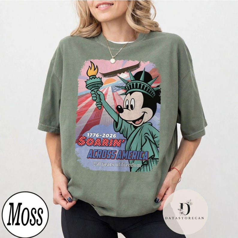 May include: Sage green t-shirt with a graphic of Mickey Mouse as the Statue of Liberty, holding a torch. The design includes the text "1776-2026 Soarin' Across America" with a patriotic red, white, and blue background. The shirt has a relaxed fit.
