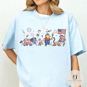 Op de afbeelding: Lichtblauw T-shirt met een patriottisch ontwerp met stripfiguren. De afbeelding bevat Winnie de Poeh, Knorretje, Iejoor en Tijger, elk versierd met Amerikaanse vlagmotieven en met een fakkel of vlag. Vuurwerk en feestelijke elementen maken het ontwerp compleet.