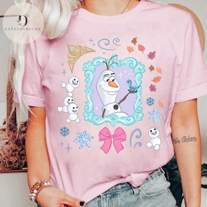 Peut inclure: T-shirt rose clair avec un dessin anim&eacute; d'Olaf de La Reine des Neiges, entour&eacute; de flocons de neige, de feuilles et d'un n&oelig;ud rose. Le motif comprend un oiseau bleu et de petits bonhommes de neige. Le t-shirt porte le logo Datastorecan.