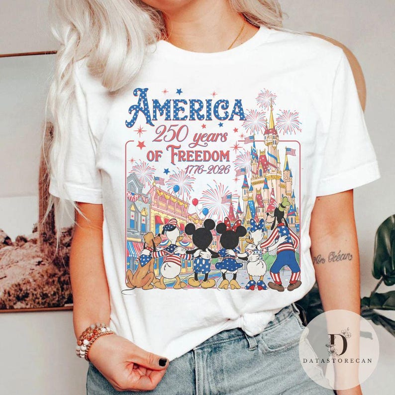May include: White t-shirt with a vibrant graphic commemorating 250 years of American freedom. The design includes fireworks, a castle, and cartoon characters. The text "America 250 years of Freedom 1776-2026" is displayed in blue.