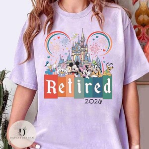 May include: A light purple t-shirt featuring a colorful graphic of Disney characters, a castle, and fireworks. The word "Retired" is displayed in large letters, with "2026" below. The design includes Mickey Mouse, Donald Duck, and other characters.