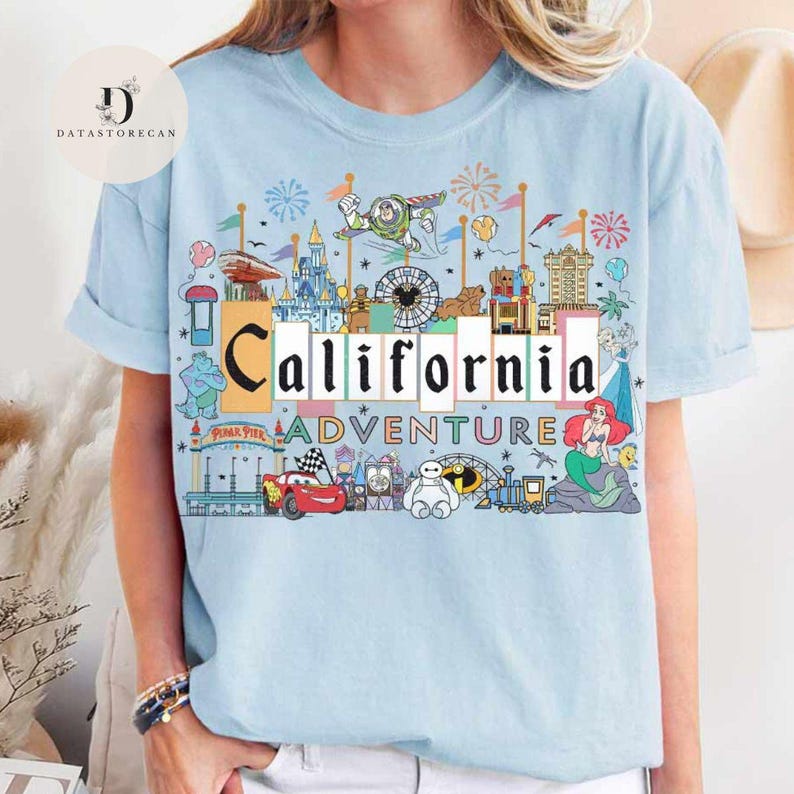 Op de afbeelding: Lichtblauw T-shirt met een kleurrijke afbeelding van het California Adventure pretpark. De afbeelding bevat iconische personages en attracties zoals Mickey Mouse, Buzz Lightyear, Ariel en de Cars.