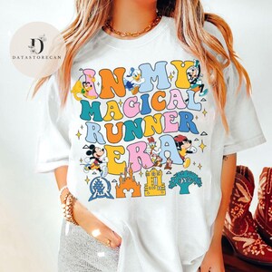 K&ouml;nnte beinhalten: Wei&szlig;es T-Shirt mit einem bunten Grafikdruck, der "In My Magical Runner Era" lautet. Die Grafik enth&auml;lt Mickey Mouse, Donald Duck und andere Disney-Figuren.