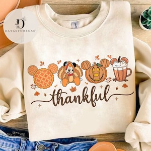 May include: Cream-colored sweatshirt featuring a Thanksgiving-themed graphic. The design includes a pie, Mickey Mouse dressed as a turkey, a pumpkin, and a coffee cup, with the word "thankful" in brown script.