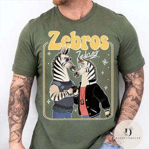 Pu&ograve; includere: T-shirt verde oliva con una grafica a cartoni animati di due zebre. Le zebre indossano occhiali da sole e giacche, con la scritta "Zebros" in un font retr&ograve; giallo. Il design &egrave; racchiuso in un rettangolo verde chiaro.