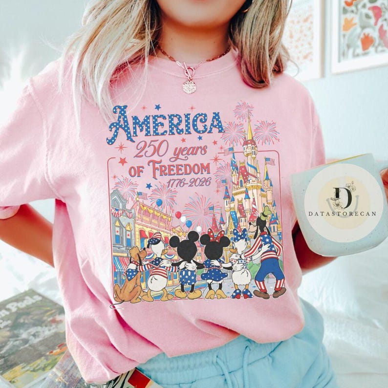 May include: A pale pink t-shirt with a graphic commemorating 250 years of American freedom. The design includes fireworks, a castle, and cartoon characters. The text reads "America 250 years of Freedom 1776-2026."