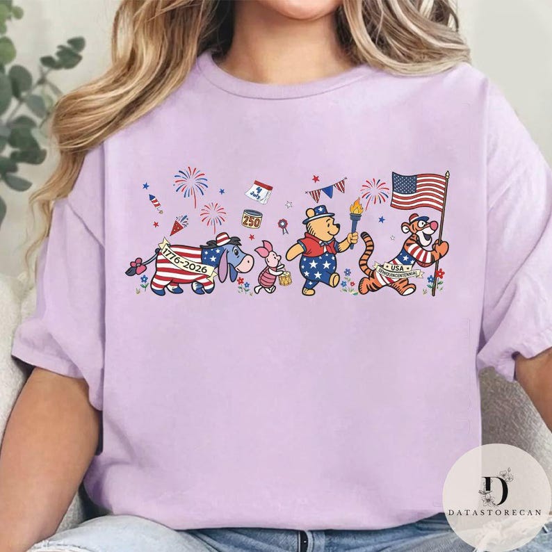 Op de afbeelding: Een lichtpaars T-shirt met een patriottisch cartoonontwerp. Het ontwerp toont een ezel, Knorretje, Winnie de Poeh en Teigetje, allemaal versierd met Amerikaanse vlagmotieven en met voorwerpen zoals een fakkel en een vlag. Vuurwerk en sterren maken de feestelijke look compleet.