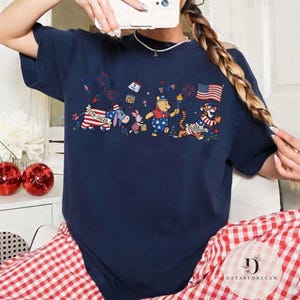 Op de afbeelding: Marineblauw T-shirt met een patriottisch ontwerp met stripfiguren. De afbeelding bevat Winnie de Poeh en Tijger, die de VS vieren met vuurwerk en vlaggen. Het ontwerp is in rood, wit en blauw.