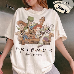 May include: An ivory t-shirt featuring a graphic print of various animated characters sitting on a couch. Below the illustration, the text reads "FRIENDS SINCE 1995". The design includes Woody, Buzz Lightyear, Jessie, Rex, Bullseye, and other characters.