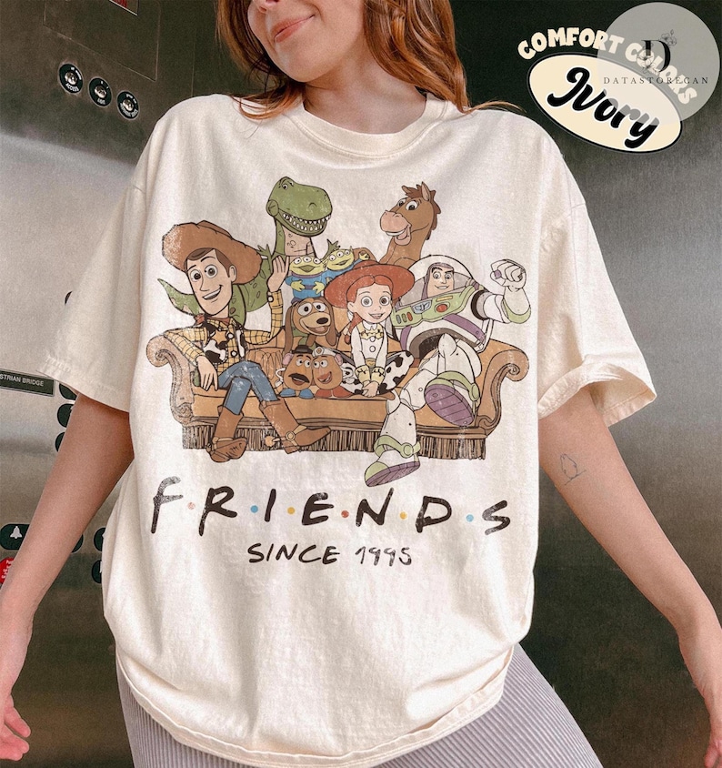 May include: An ivory t-shirt featuring a graphic print of various animated characters sitting on a couch. Below the illustration, the text reads "FRIENDS SINCE 1995". The design includes Woody, Buzz Lightyear, Jessie, Rex, Bullseye, and other characters.