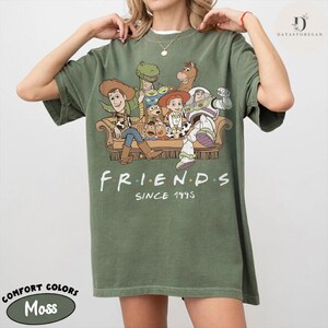 May include: An olive green short-sleeve t-shirt featuring a graphic print of various animated characters sitting on a couch. Below the characters, the text reads "FRIENDS" and "SINCE 1995". The shirt has a crew neckline and a relaxed fit.