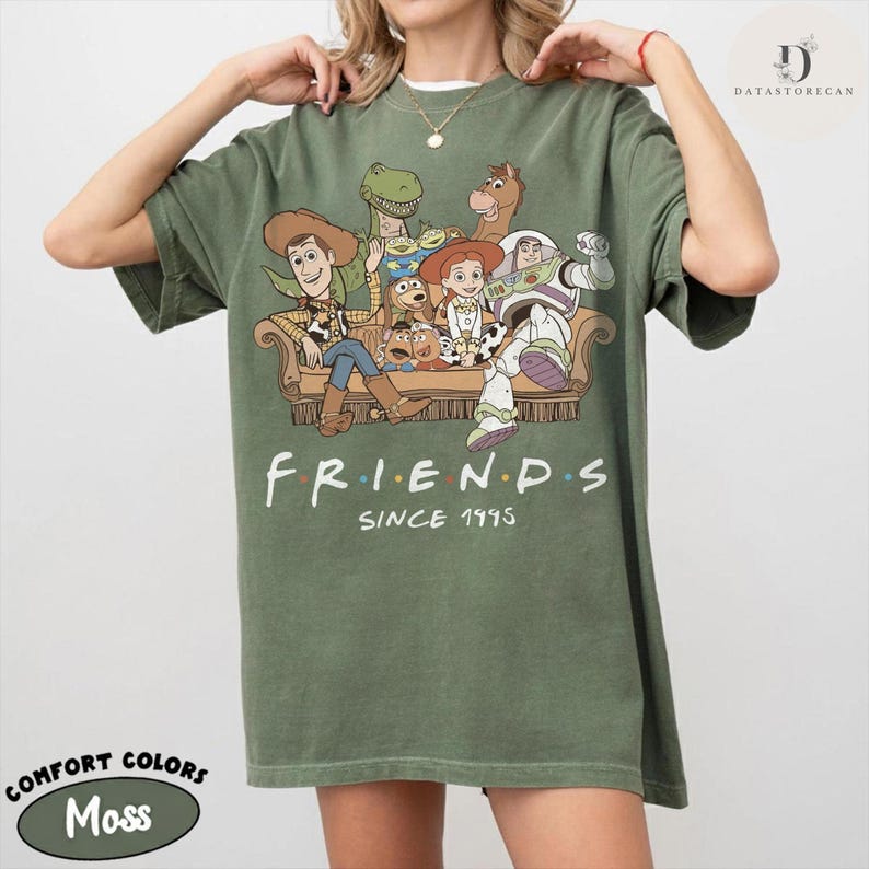 May include: An olive green short-sleeve t-shirt featuring a graphic print of various animated characters sitting on a couch. Below the characters, the text reads "FRIENDS" and "SINCE 1995". The shirt has a crew neckline and a relaxed fit.