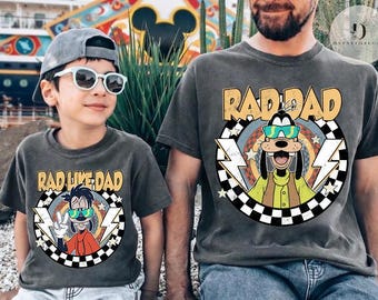 May include: Two matching charcoal gray t-shirts. One says "RAD DAD" with a cartoon Goofy wearing sunglasses. The other says "RAD LIKE DAD" with a similar Goofy graphic. Both shirts have a checkered border and lightning bolts.